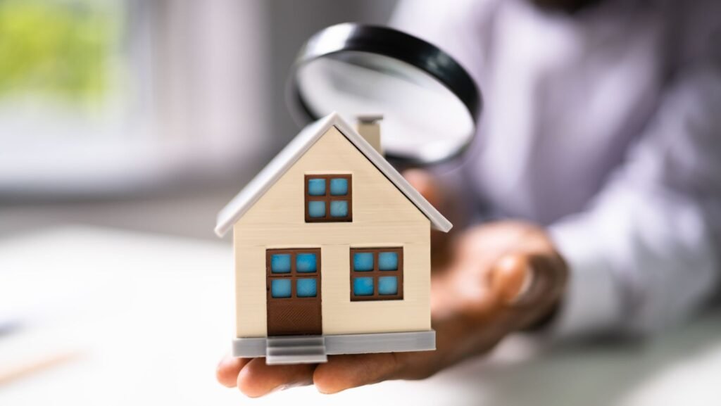 Person inspecting a small model house with a magnifying glass, symbolizing a home inspection.