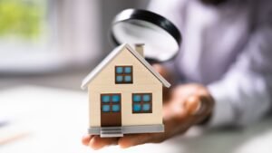 Person inspecting a small model house with a magnifying glass, symbolizing a home inspection.
