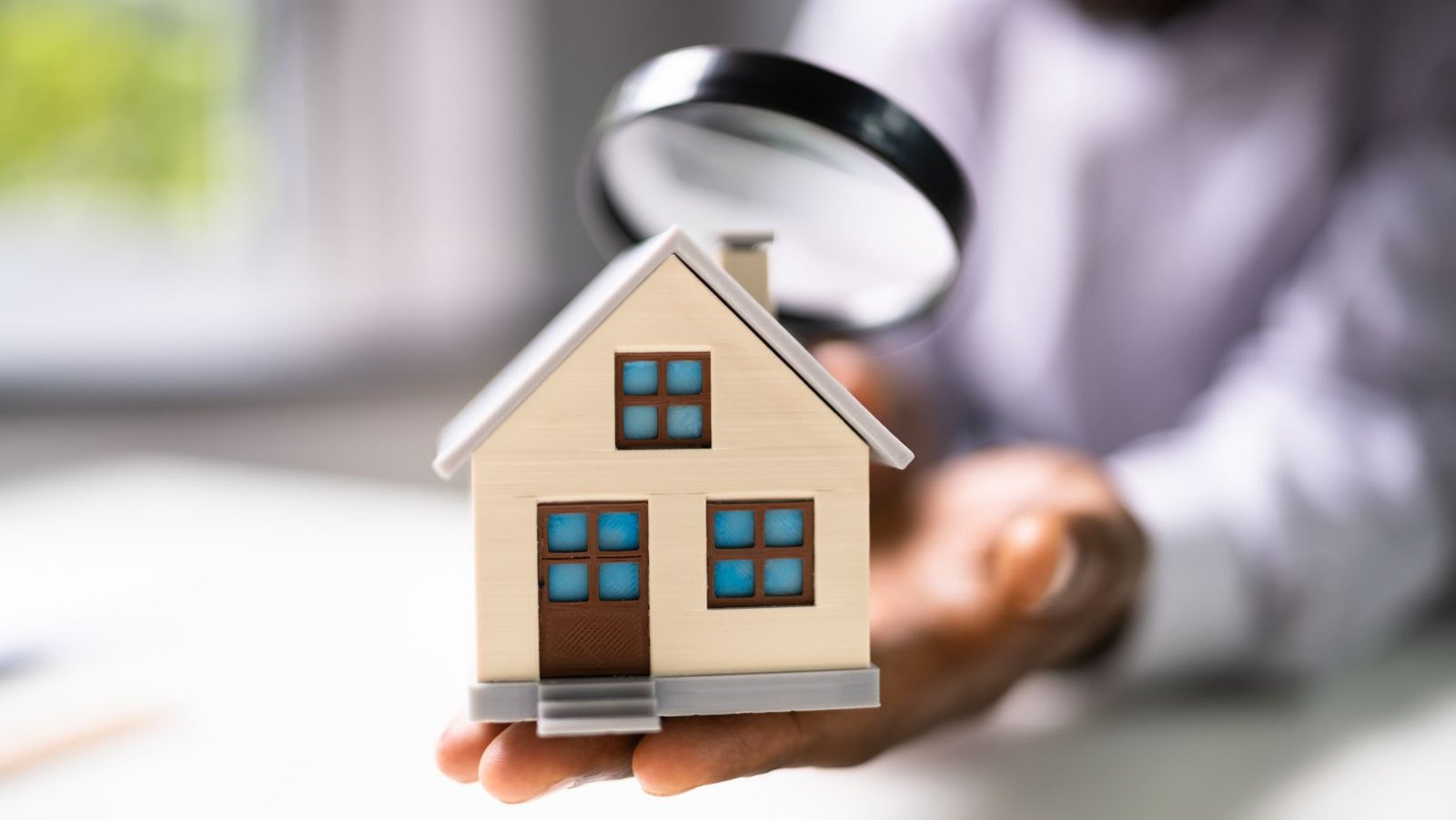 Person inspecting a small model house with a magnifying glass, symbolizing a home inspection.